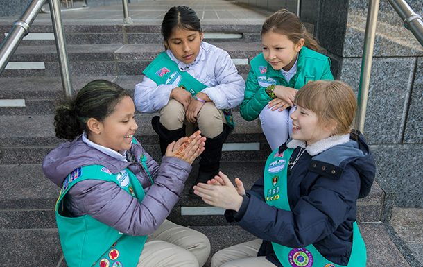 Four girl scouts in uniform sitting outside on steps playing a game.